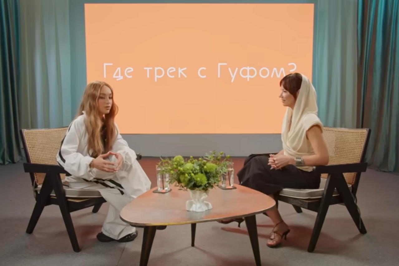 Арендовать А53: Secret Gallery (Секретная галерея). Современное студийное пространство с двумя плетеными креслами, овальным деревянным столиком, двумя микрофонами и растением в вазе. На заднем плане экран с надписью: "Где трек с Гуфом?". Шторы в светло-бирюзовых тонах.