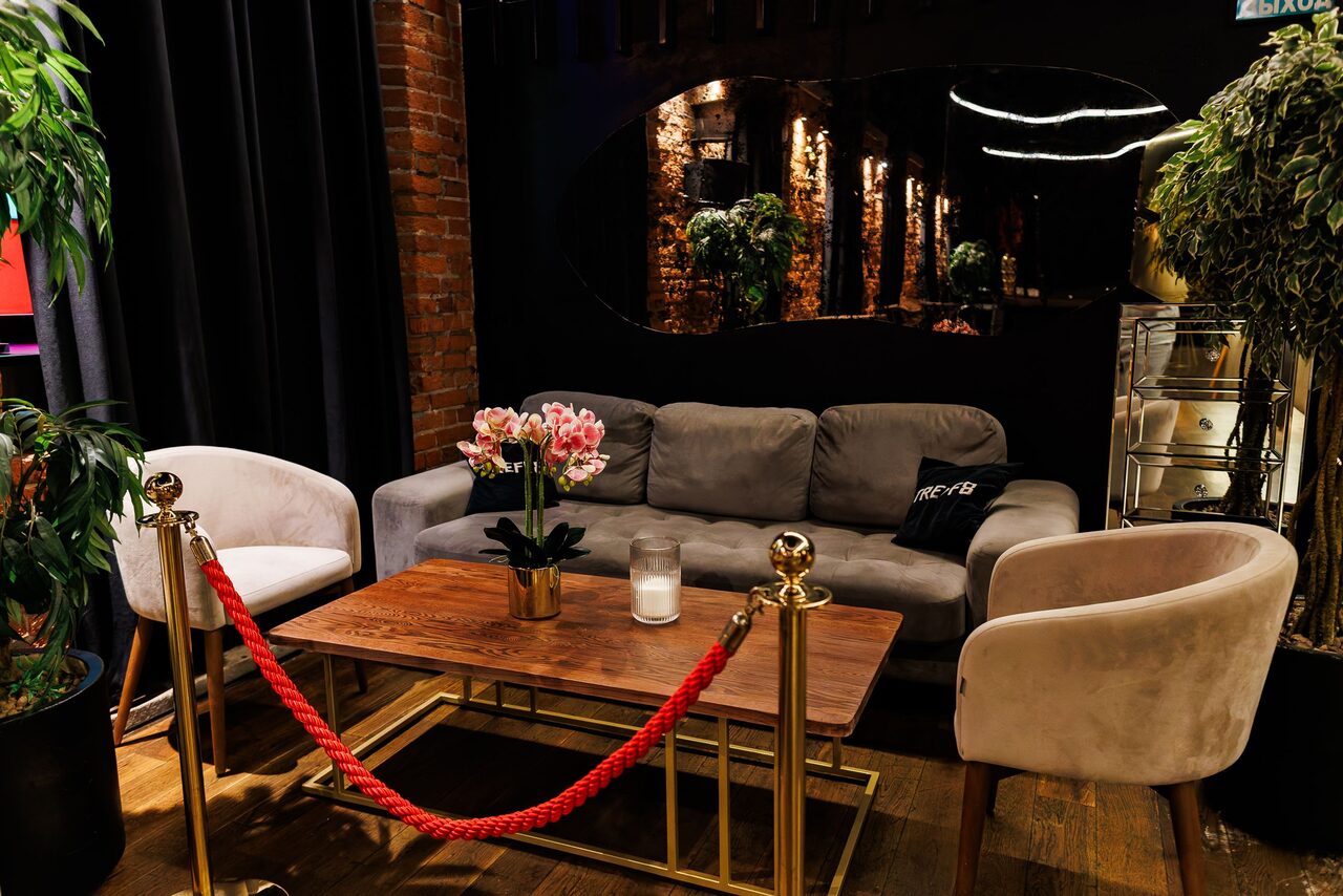 Арендовать Двухуровневая event-площадка 550 м². Лаунж пространство с диваном и двумя креслами вокруг деревянного стола на золотых ножках. Красный канат на стойках создает барьер. На столе свеча и цветы в горшке. На стене темное зеркало. Кирпичная стена и деревянный пол. Тёмные шторы слева.