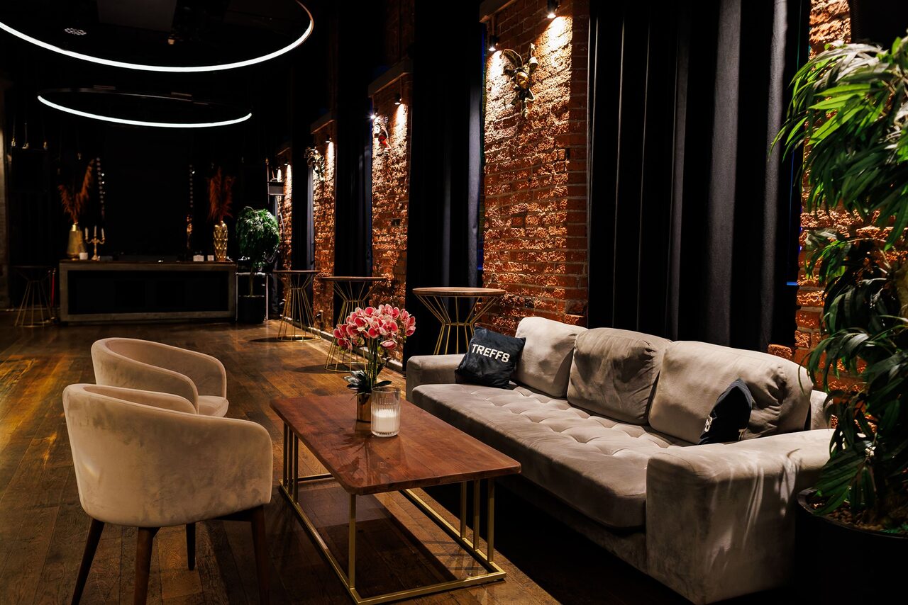 Арендовать Двухуровневая event-площадка 550 м². Интерьер просторного помещения с кирпичными стенами и деревянным полом. Зона отдыха с серым диваном, двумя бежевыми креслами, деревянным столом и декором. Потолочные светильники в виде кругов, текстиль тёмных тонов, зелёные растения.