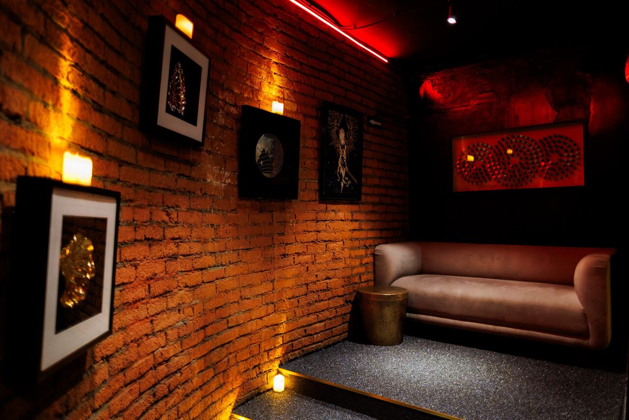 Арендовать Двухуровневая event-площадка 550 м². Небольшое помещение с кирпичными стенами, украшенными картинами в рамках. Мягкий диван и металлический столик. Пол устелен ковровым покрытием. Освещение от потолочных светильников и подсветка на стенах. Цвета: коричневый, черный, красный.