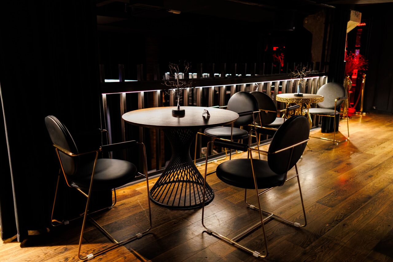 Арендовать Двухуровневая event-площадка 550 м². Пространство с деревянным полом и круглыми столиками. Рядом черные стулья с металлическими элементами. На фоне барная стойка с вертикальными полосами и подсветкой. Освещение приглушенное, элементы декора в черных и золотистых тонах.