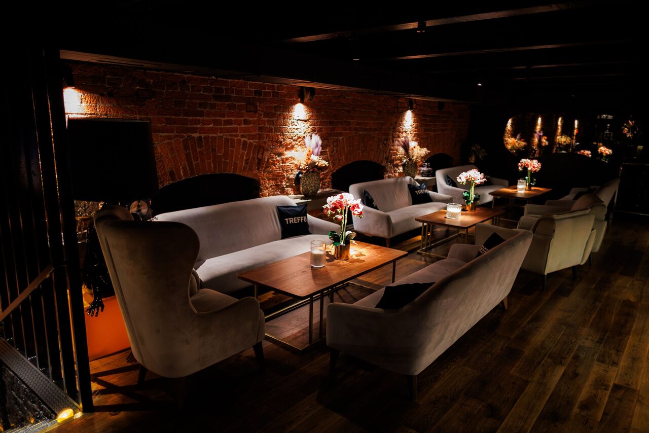 Арендовать Двухуровневая event-площадка 550 м². Лофт-пространство с кирпичной стеной и деревянным полом. Мягкие диваны и кресла расположены вокруг деревянных столов. Настенные светильники, расставленные цветы на столах. Интимное освещение, зеркала на стене.