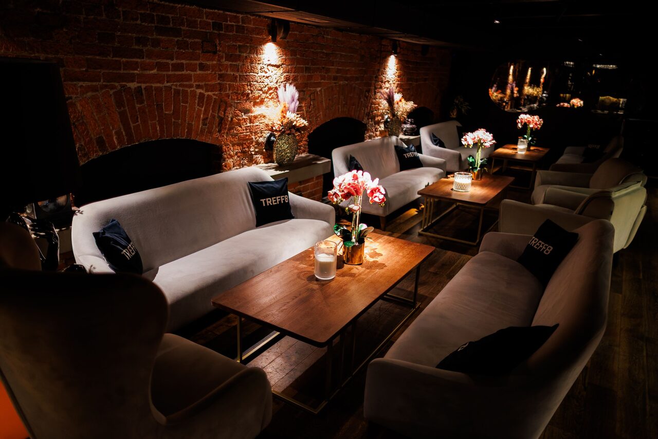 Арендовать Двухуровневая event-площадка 550 м². Лаунж-пространство с кирпичными стенами и мягким освещением. Расставлены серые диваны и кресла, деревянные столы с цветами и свечами. Пары декоративных вазы на столах. Тёмный пол, подушки на диванах. Приглушённый уютный интерьер.