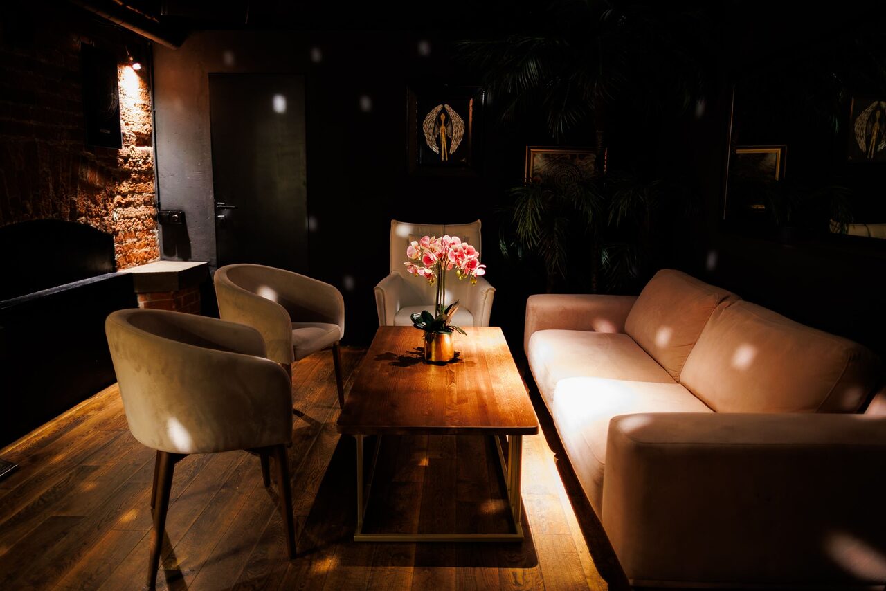 Арендовать Двухуровневая event-площадка 550 м². Мягкая зона отдыха с диваном, двумя креслами и креслом с высокой спинкой вокруг деревянного стола. На столе ваза с цветами. Настенный светильник с теплым освещением. Стены из кирпича и темные. Темное деревянное напольное покрытие.
