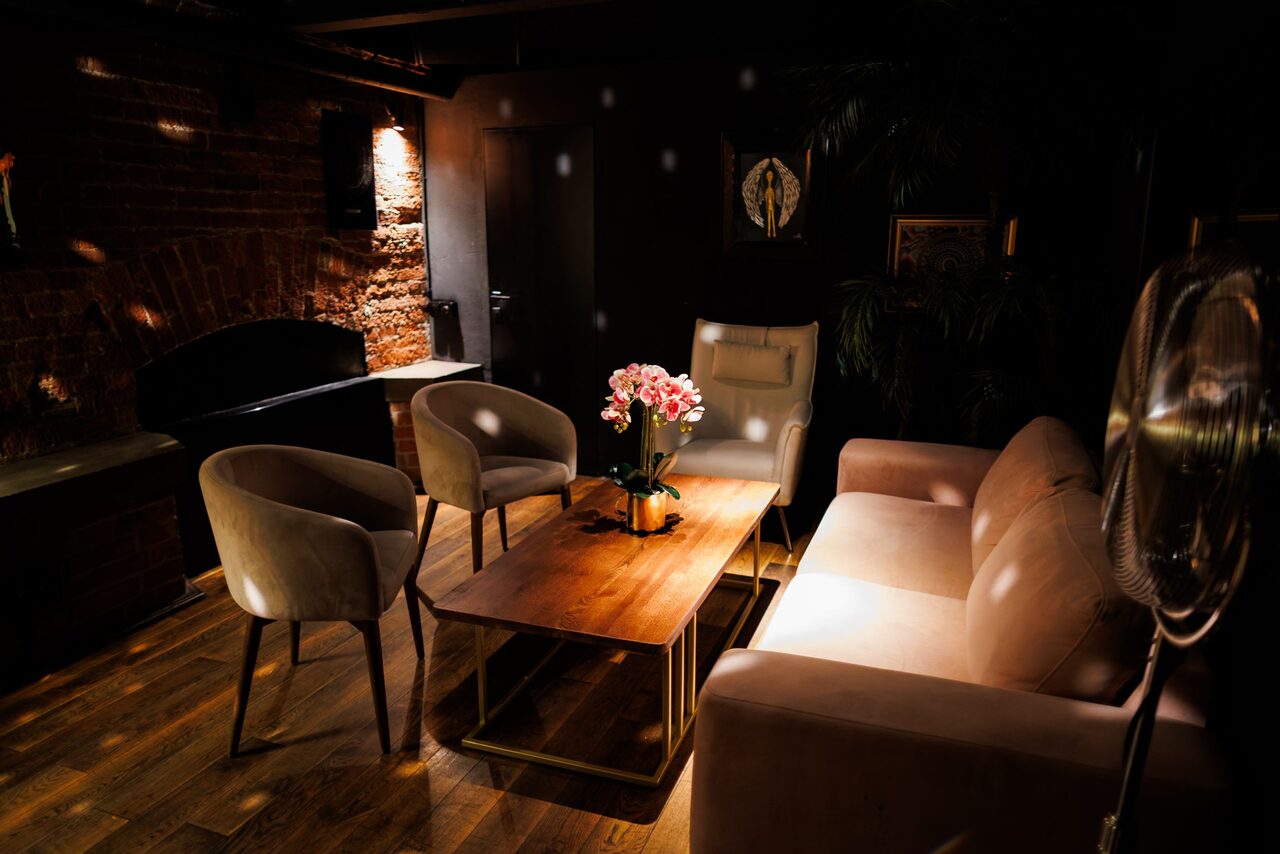 Арендовать Двухуровневая event-площадка 550 м². Интерьер комнаты с кирпичной стеной и деревянным полом. В центре кофейный столик с розовыми цветами, окруженный мягким диваном и креслами. Теплое освещение и декоративный камин. На стене висят картины и зеркало.