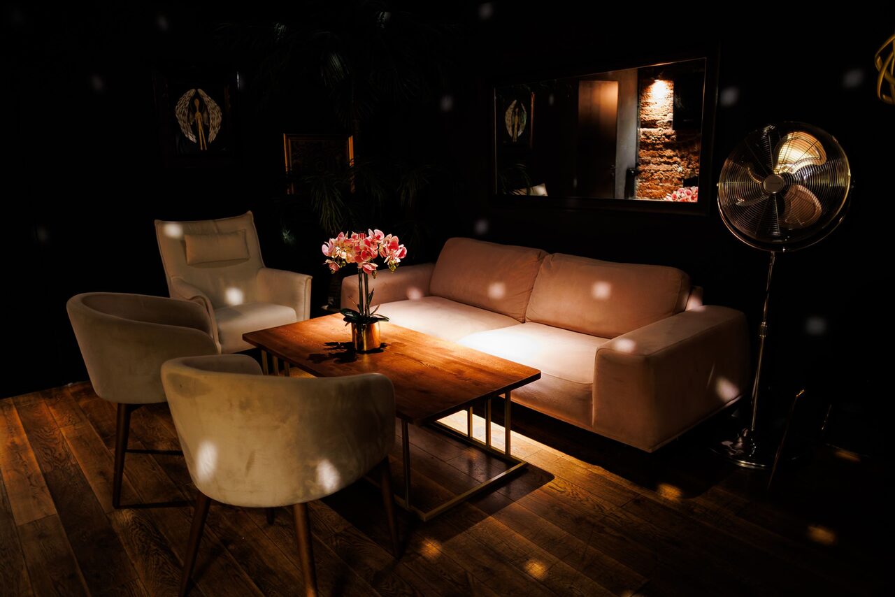 Арендовать Двухуровневая event-площадка 550 м². Интерьерная зона с мягким диваном и креслами вокруг деревянного стола, на котором стоят цветы. Потолочное освещение создает точки света по помещению. Напольное покрытие из темного дерева. Слева кресло, справа вентилятор, на стене зеркало.