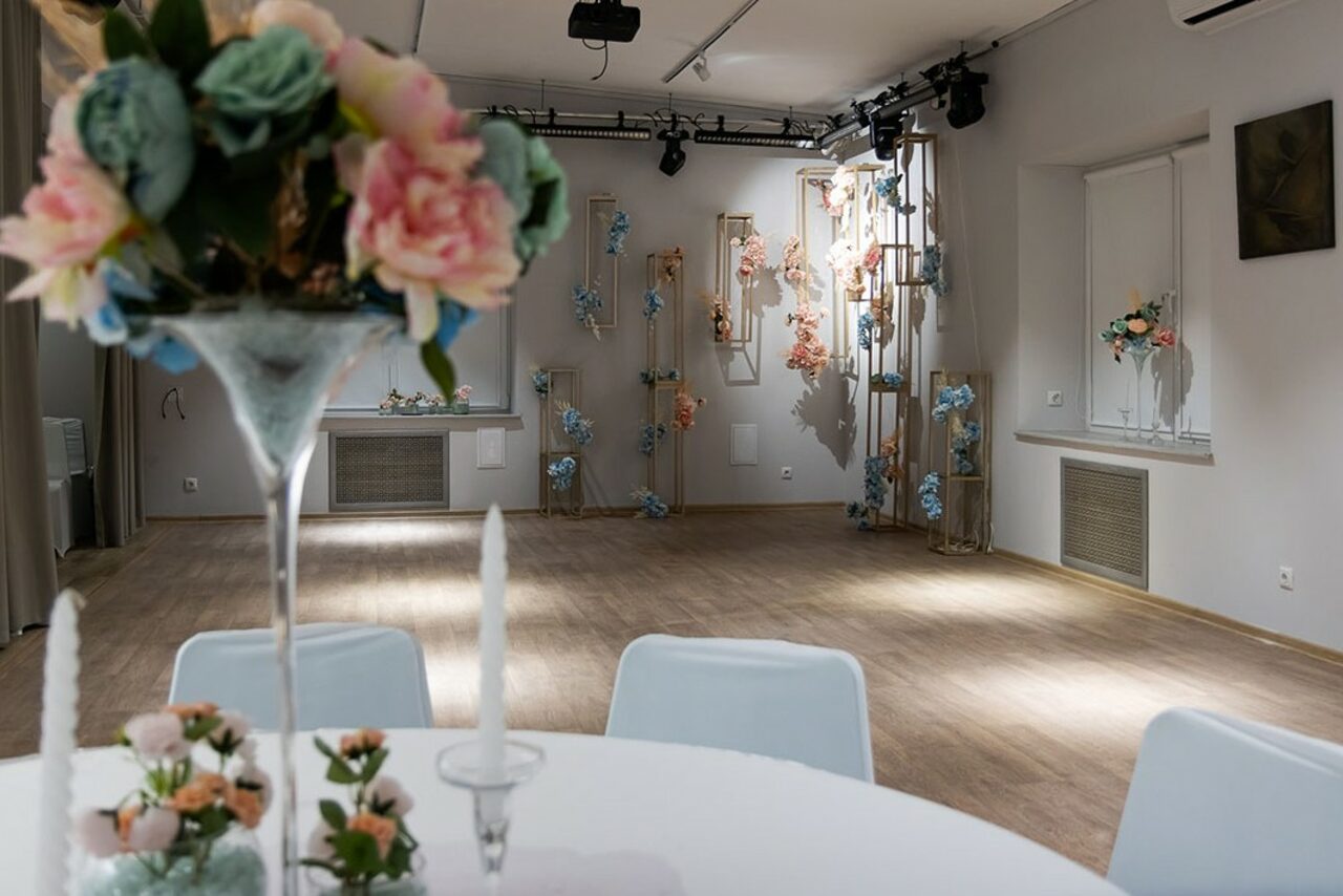 Арендовать Пространство для ваших праздников. Просторное помещение с деревянным полом, белыми стенами, круглым столом с белой скатертью, цветами и свечами. На стенах и стойках декоративные цветочные композиции. Освещение точечное, присутствует оборудование для световых эффектов.