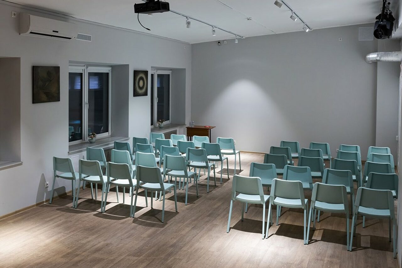 Арендовать Пространство для ваших праздников. Просторная комната с серыми стенами и деревянным полом, расставленные в ряды голубые пластиковые стулья, два окна с подоконниками, потолочный регулированный свет, настенные декоративные элементы, кондиционер, вытяжка, небольшой деревянный стол.