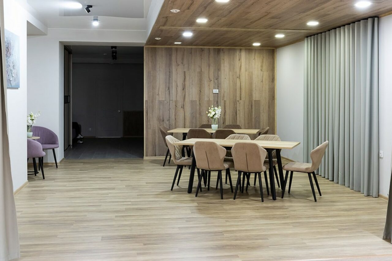 Арендовать Пространство для ваших праздников. Современное помещение с деревянным полом и стенами, двумя столами и десятью стульями с бежевой обивкой. Мягкое потолочное освещение, серые занавески, декоративные цветы на столах. Спокойные нейтральные цвета.