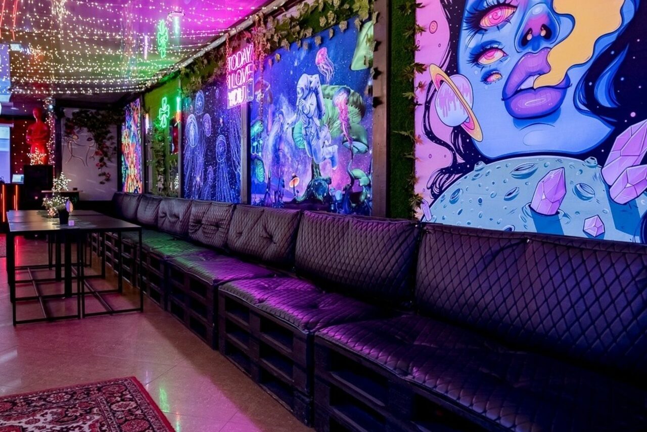 Арендовать Яркий лофт для праздников. Лофт-пространство с длинным деревянным столом и черными кожаными диванами. Яркие граффити на стенах. Подвесные гирлянды разноцветных ламп. Напольное покрытие из светлого ламината. Зеленые декоративные растения вдоль стен.