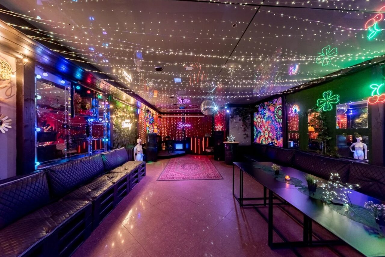 Арендовать Яркий лофт для праздников. Просторное помещение с длинными диванами вдоль стен, низкими столами и цветными неоновыми украшениями на стенах. Потолок украшен множеством маленьких светодиодов. Пол гладкий, с розовым ковром. Зеркала на стенах, приглушенное освещение.