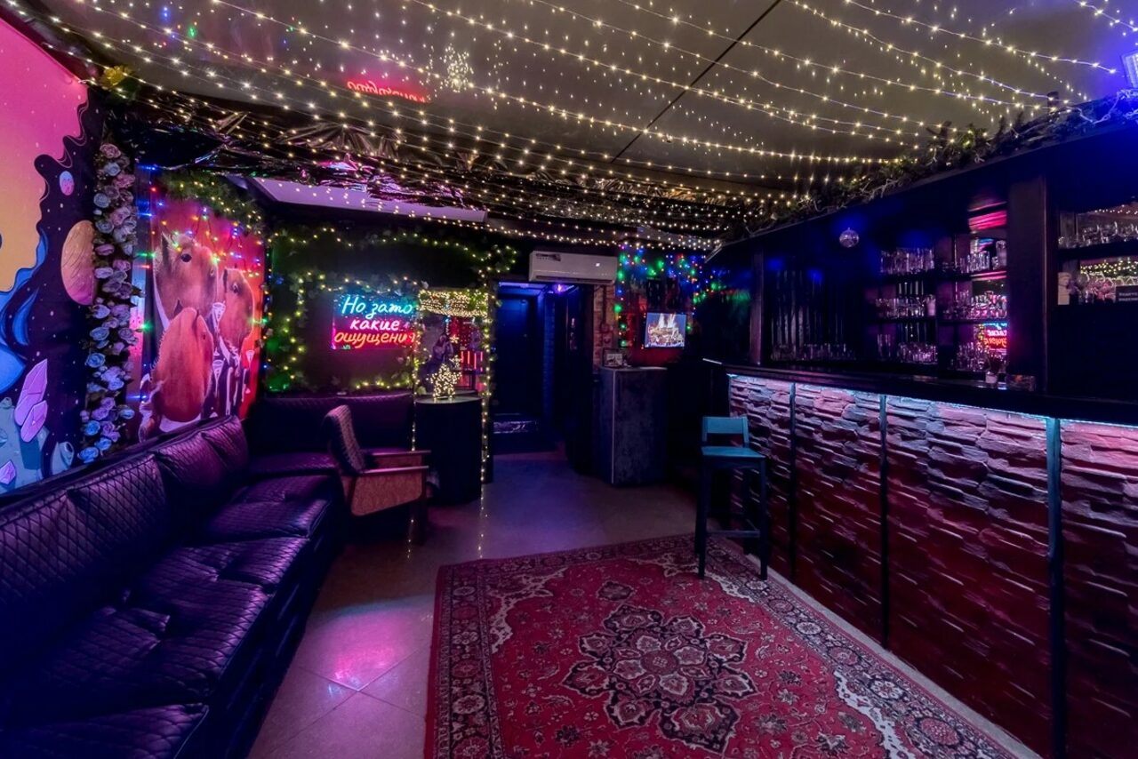 Арендовать Яркий лофт для праздников. Лаунж-пространство с барной стойкой, черной кожаной мебелью и деревянными элементами отделки. Потолок украшен гирляндами. На стене яркие граффити и неоновая вывеска. Ковровое покрытие с орнаментом на полу.