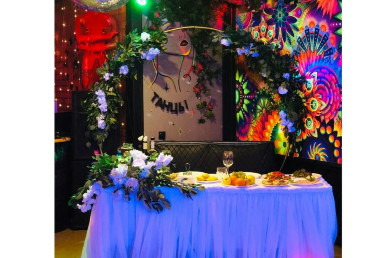 Арендовать Яркий лофт для праздников. Пространство с праздничным декором, длинный стол с белой скатертью и блюдами, арка из зелени и белых цветов. На стене яркий абстрактный узор и постер с надписью "Танцы". Разноцветное освещение, стул с подушкой, блестящая занавеска.