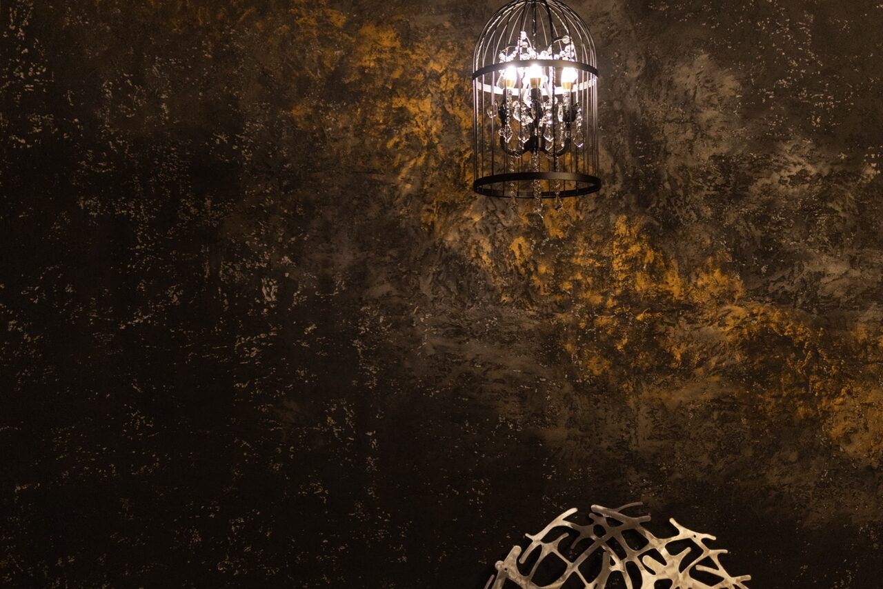 Арендовать Лофт со своим двориком. Темная стена с текстурой и золотистыми акцентами, подвесной светильник в форме клетки с лампой внутри, металлический декоративный элемент на переднем плане. Освещение теплое и направленное.