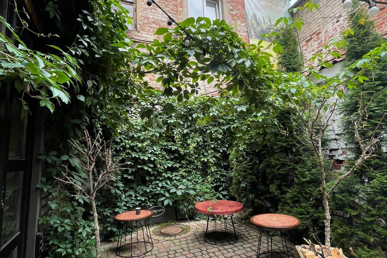 Арендовать Лофт со своим двориком. Уютный внутренний двор с кирпичными стенами, покрытыми зеленью. Три круглых деревянных стола на металлических ножках. Каменная мостовая с растущими вокруг кустами и деревьями. Верхнее естественное освещение.