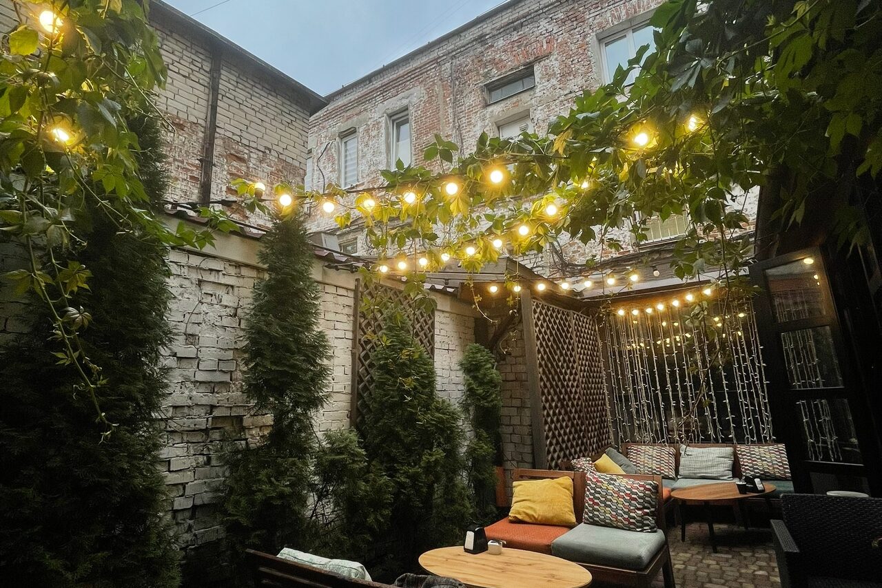 Арендовать Лофт со своим двориком. Уличное пространство с кирпичными стенами, выложенной плиткой площадкой и диванами с подушками. Множество растений и гирлянд с лампочками. Деревянные круглые столы, мягкие сиденья, разноцветные подушки. Вертикальные решетки с растениями.