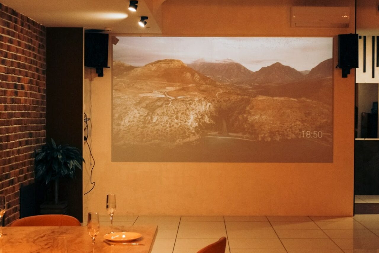 Арендовать Антикафе с несколькими залами. Помещение с кирпичной стеной слева и гладкой стеной, на которой проецируется изображение горного пейзажа. Пол с крупной плиткой. Стол из дерева с двумя бокалами и пустой тарелкой. Потолочные светильники обеспечивают мягкое освещение.