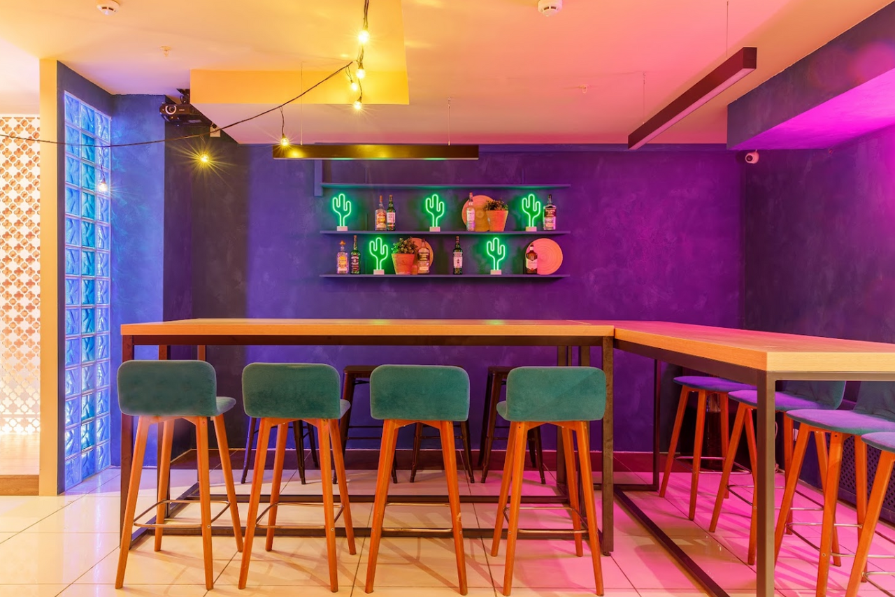 Арендовать Антикафе с несколькими залами. Барное пространство с высоким столом и стульями с зелеными сиденьями. Фиолетовая стена с полкой, на которой расположены неоновые фигуры кактусов и бутылки. Яркое освещение с теплым и фиолетовым оттенком, плиточный пол.