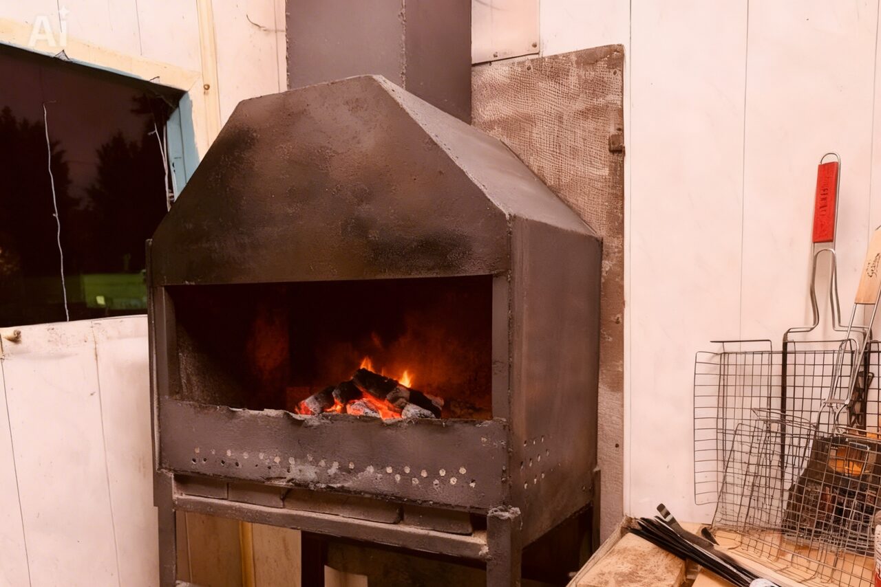 Арендовать Лофт-мансарда . Камера с металлическим камином или мангалом, в углу великая металлическая решетка и инструменты для барбекю. Стены выкрашены в светлый цвет, у окна видна тёмная занавеска. Справа на деревянной поверхности размещена ёмкость.