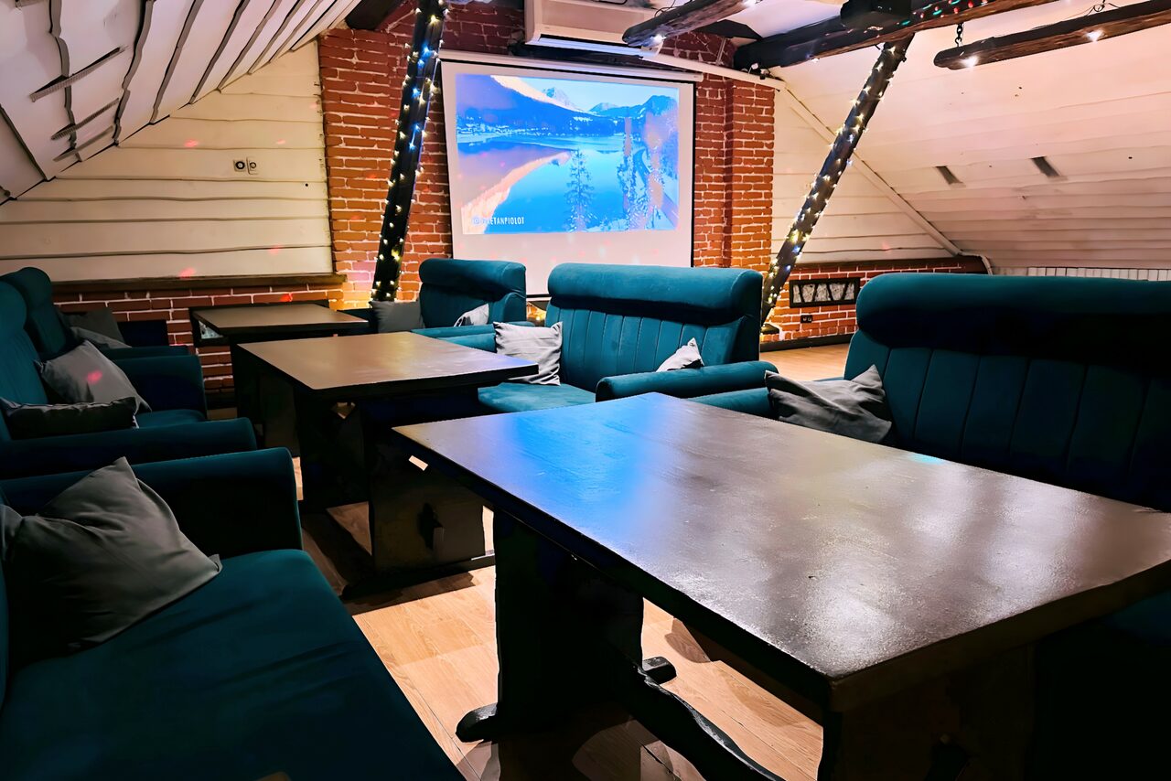 Арендовать Лофт-мансарда . Мансарда с кирпичными стенами и деревянными панелями, с синими мягкими диванами, рядом несколько прямоугольных столов. Большой экран проектора на фоне. Потолок с черными балками. Теплое освещение, гирлянды на стойках. Пол светлый деревянный.