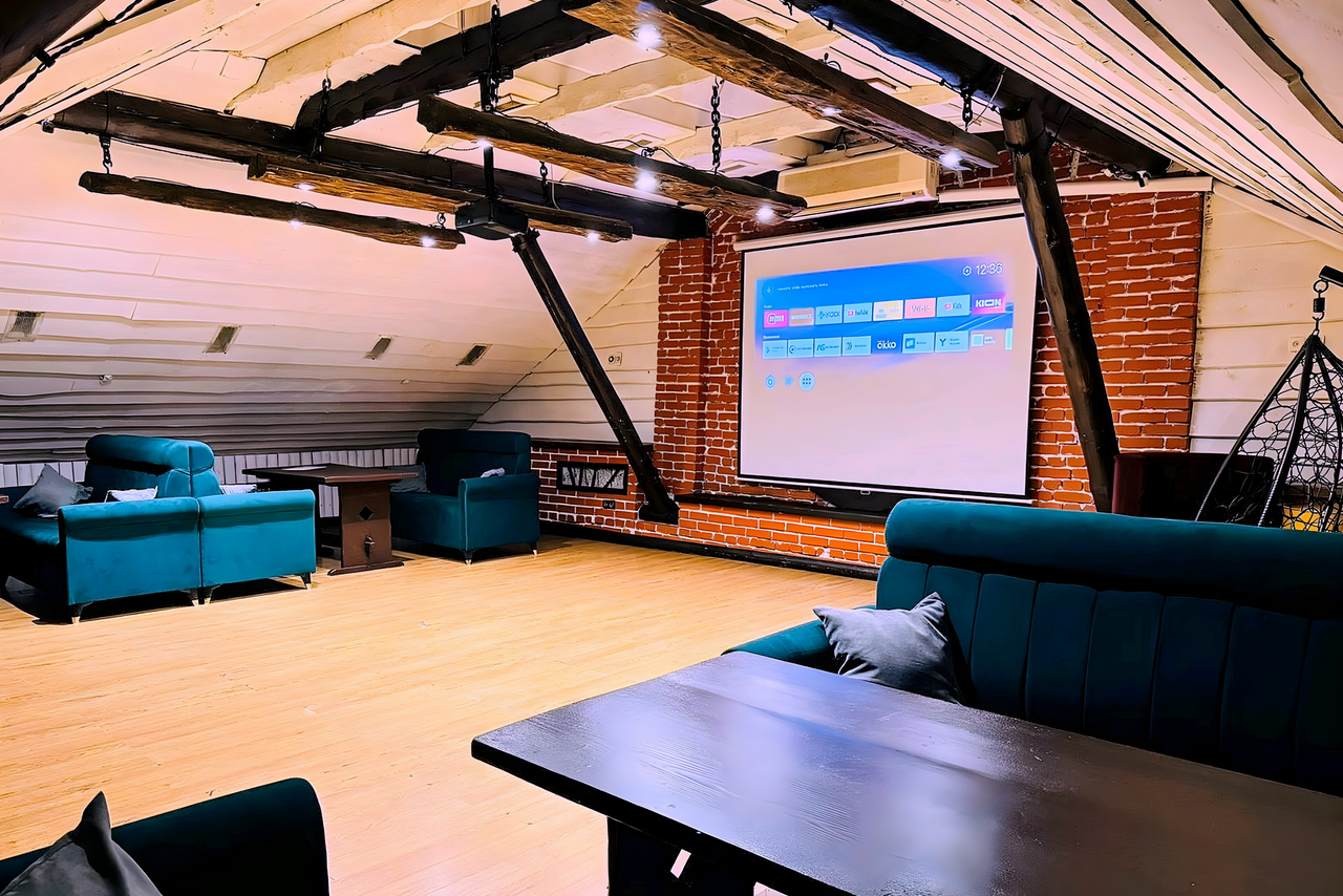 Арендовать Лофт-мансарда . Лофт-пространство с деревянным полом, кирпичной стеной и деревянными балками. Установлен проектор. Есть синие диваны, черные столы и кресла. Освещение точечными лампами. Стены окрашены в белый и серый цвета.