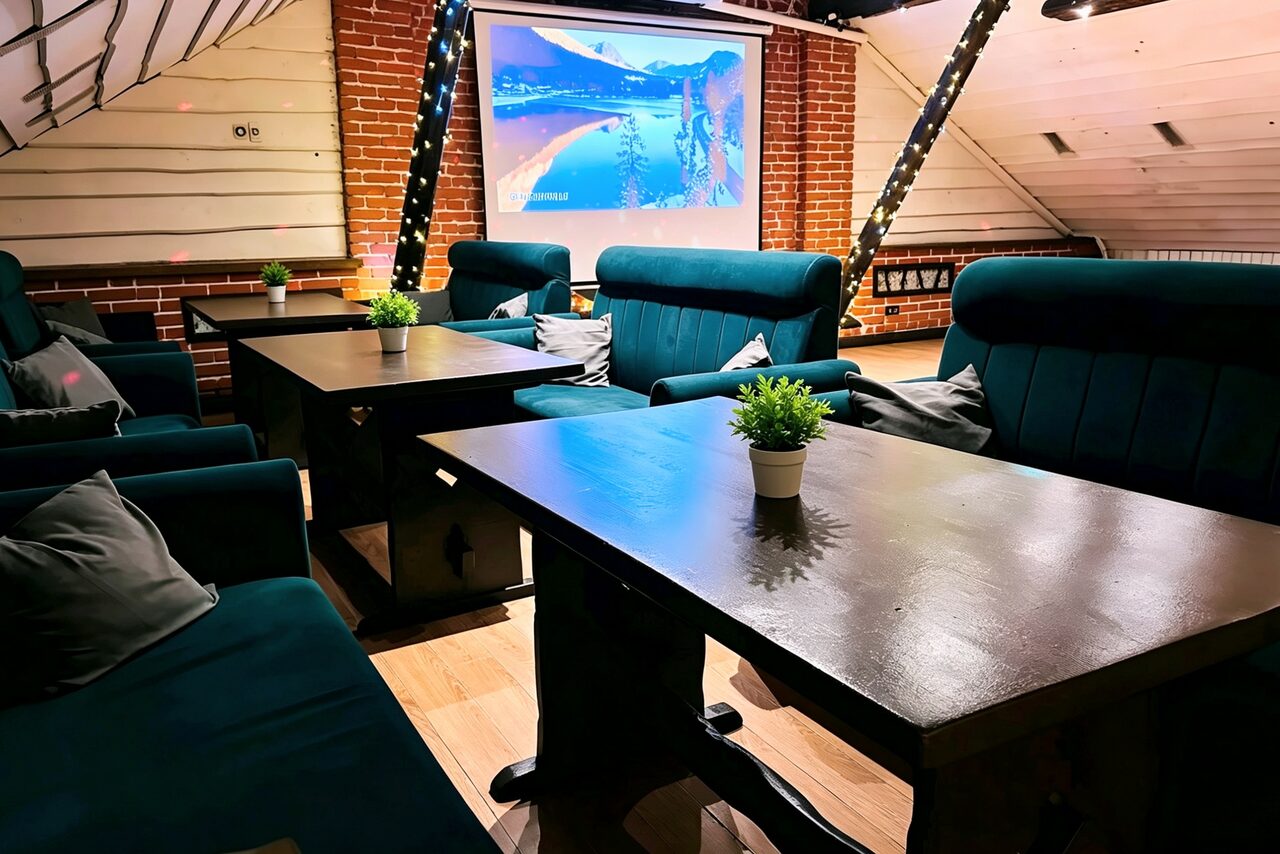 Арендовать Лофт-мансарда . Кофейня с деревянными панелями и кирпичной стеной. Темные диваны и столы. На столах зеленые растения в горшках. На стене проектор с изображением природы. Потолок с косыми линиями обрамлен гирляндами. Светлый деревянный пол.