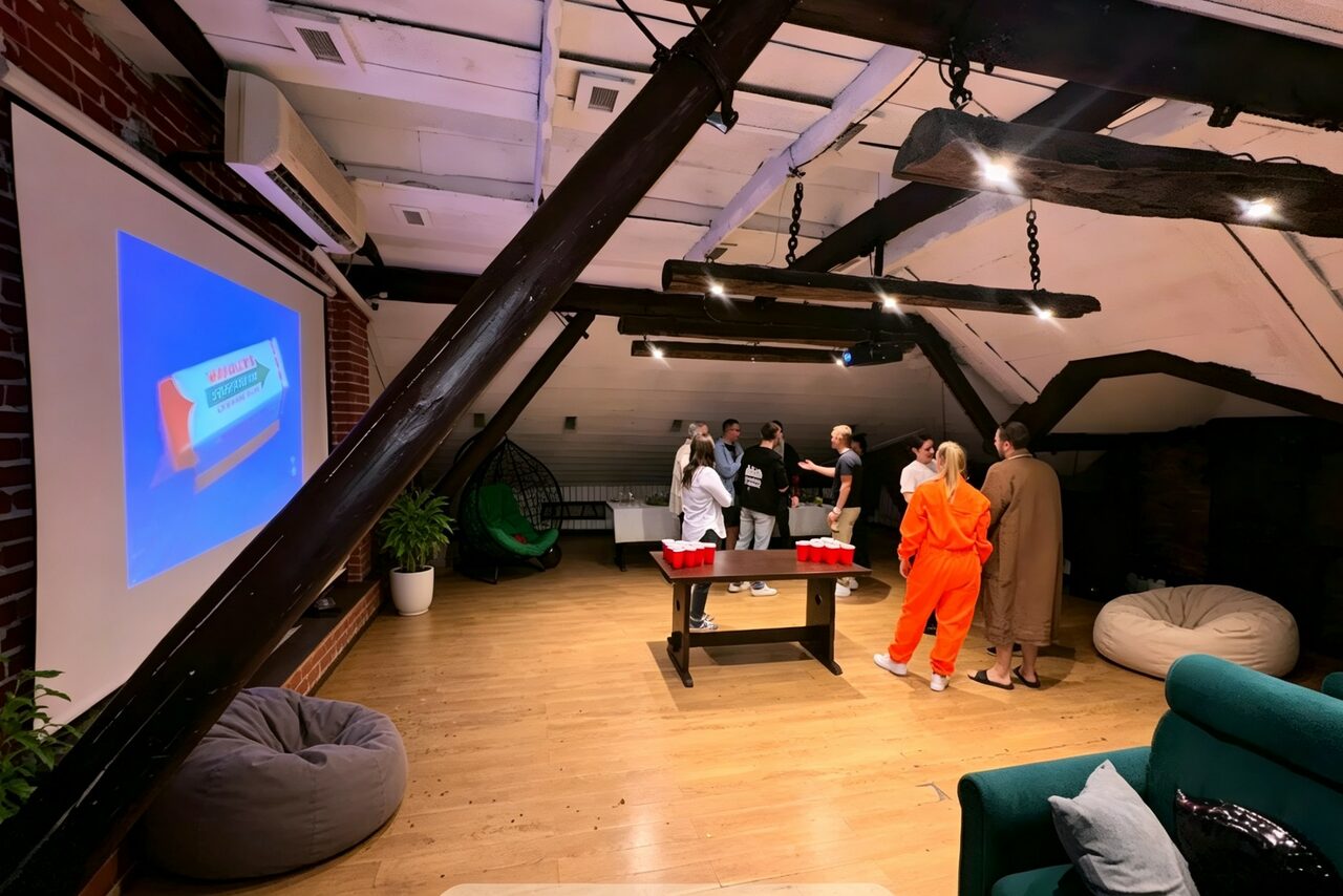 Арендовать Лофт-мансарда . Лофт-пространство с деревянными полами и кирпичной стеной. На стене проекционный экран, на полу пуфы и столик с красными стаканами. Пучковая лампа, деревянные балки, зеленый диван с подушками, группа людей.