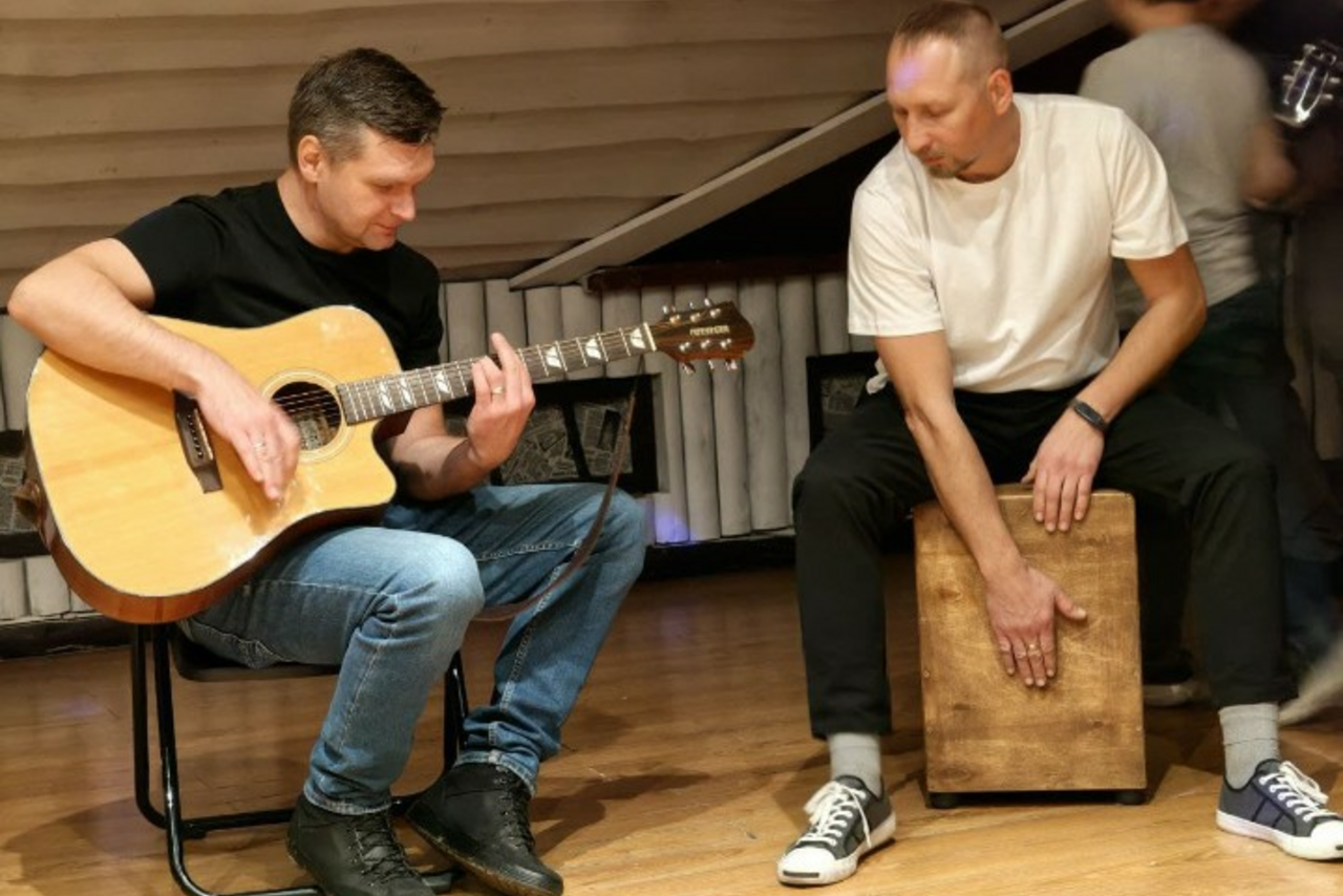 Арендовать Лофт-мансарда . Атмосферное пространство с деревянным полом и наклонным потолком с деревянной отделкой. Два человека играют на гитаре и кахоне. Один сидит на стуле, другой — на кахоне. В помещении тёплое освещение.