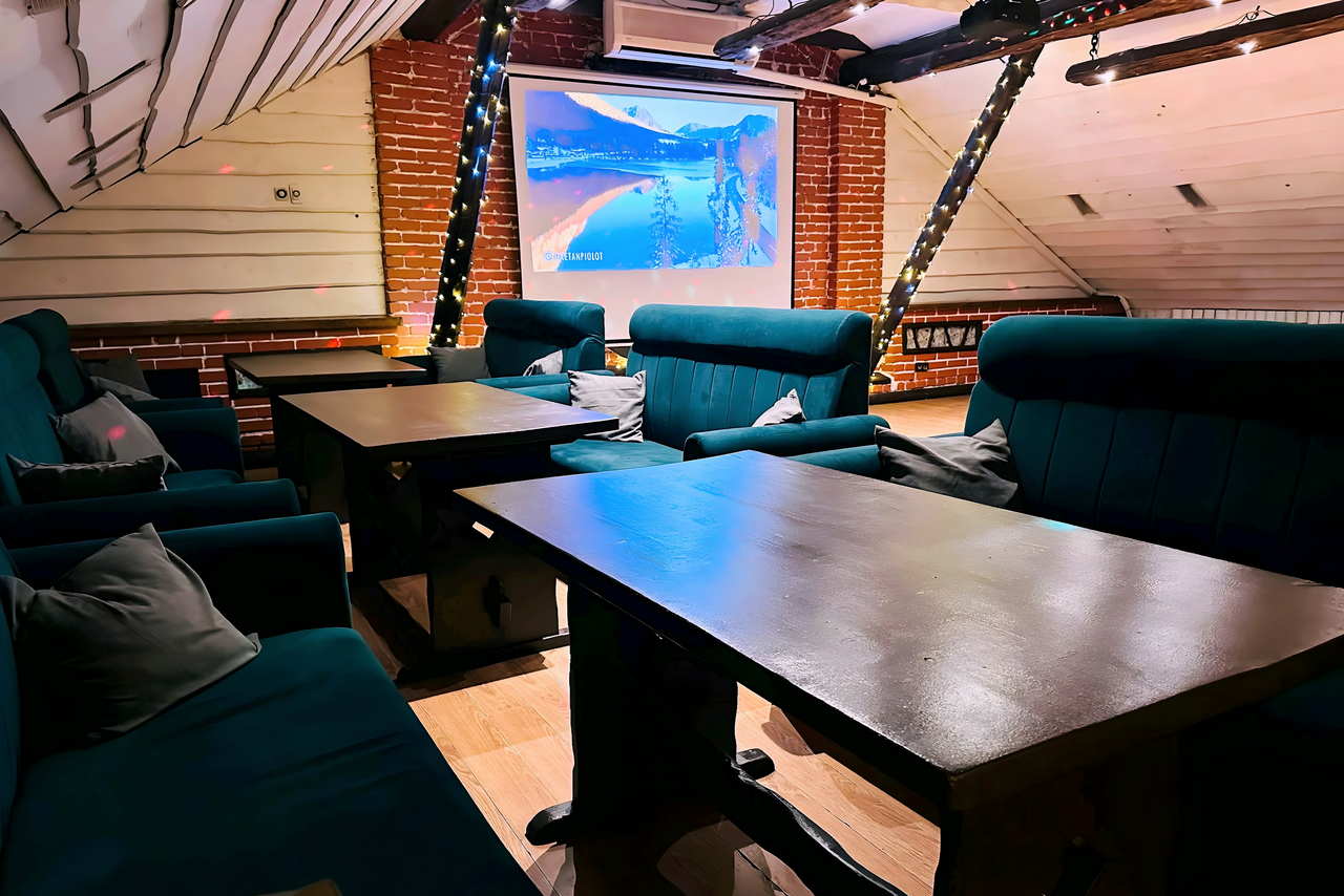 Арендовать Лофт-мансарда . Лофт-пространство с деревянными столами и темными диванами. Красный кирпич и деревянные панели на стенах. Проектор с изображением на экране. Деревянный пол, обшитый потолок. Ограниченное освещение с гирляндами. Несколько подушек на диванах.