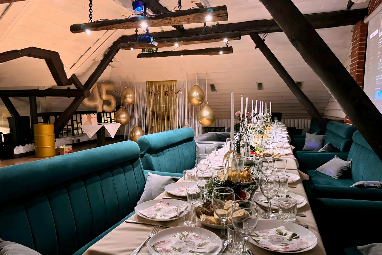 Арендовать Krisha loft (Крыша Лофт). Чердак с деревянными балками, длинный стол с белыми скатертями, сервировка тарелками и бокалами. С каждой стороны стола зелёные диваны, декоративные золотые шары, свечи, проектор, светлая стена, элементы кирпичной кладки.