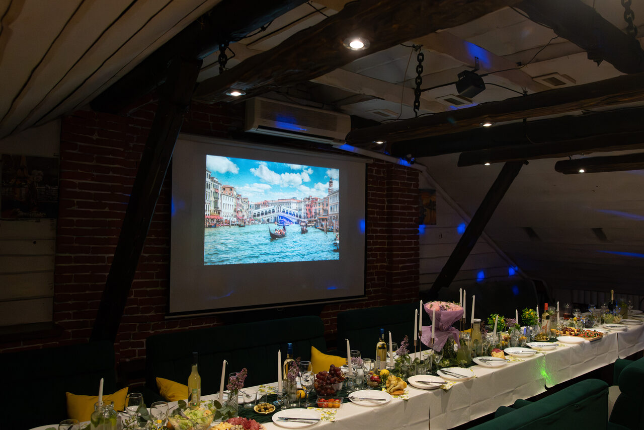 Арендовать Krisha loft (Крыша Лофт). Атмосферное помещение с деревянными балками и кирпичными стенами. Длинный стол покрыт белой скатертью, на нем расставлены тарелки, бокалы, свечи и разнообразные блюда. На стене проекционный экран с изображением города на воде. Потолочные светильники обеспечивают мягкое освещение.