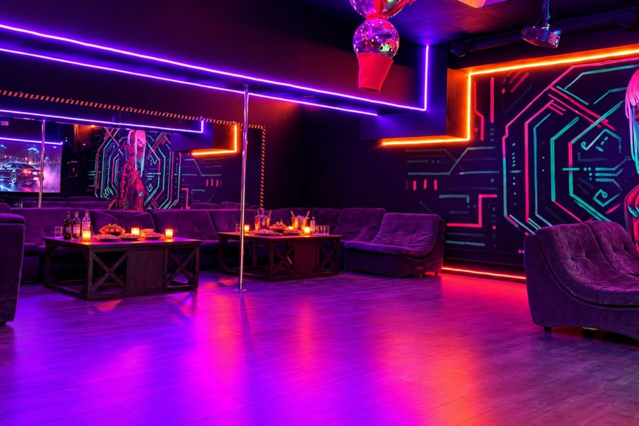 Арендовать Cyberpunk (Киберпанк). Темное помещение с неоновым освещением. По периметру мягкие диваны. На стенах граффити в стиле киберпанк. В центре металлический шест. Столы с напитками и свечами. Зеркала на стенах. Пол – деревянный. Атмосферное освещение.