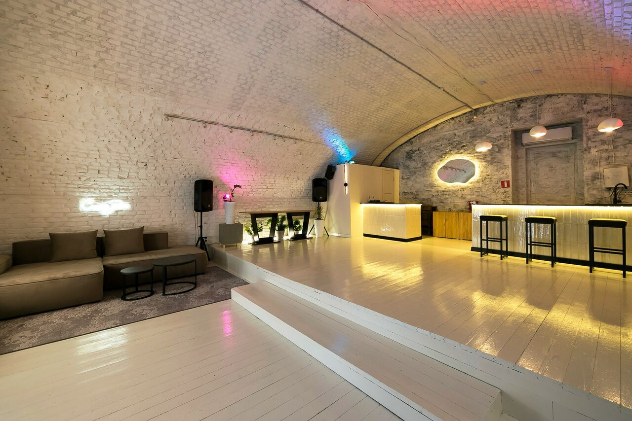 Арендовать Brodniki Loft (Бродники Лофт). Лофт из белого кирпича. Лофт с белым кирпичным арочным потолком, светлый деревянный пол, зона отдыха с диваном и стульями, барная стойка с высокими стульями, разноцветная подсветка, аудиооборудование, небольшое зеркало на стене, светлые и серые оттенки.