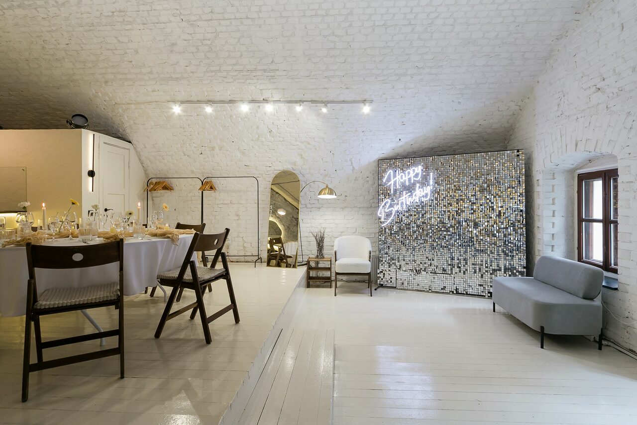 Арендовать Brodniki Loft (Бродники Лофт). Лофт из белого кирпича. Арендуемое пространство с белыми кирпичными стенами и деревянным полом. Зона с накрытыми столами и стульями, светло-серый диван и зеркало. Сверху точечные светильники. На стене светящаяся надпись "Happy Birthday".