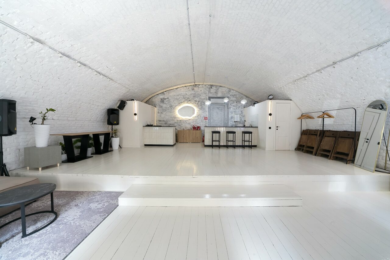 Арендовать Brodniki Loft (Бродники Лофт). Лофт из белого кирпича. Арочное пространство с белым кирпичным сводом. В центре сцена с барной стойкой и стульями. Черно-белая мебель, зеркала и аудиосистема. Деревянный пол и вешалки справа. Светлая цветовая гамма, хорошее освещение.