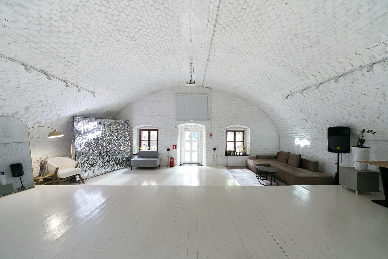 Арендовать Brodniki Loft (Бродники Лофт). Лофт из белого кирпича. Арочное помещение с белыми кирпичными стенами и деревянным полом. В центре диваны, кресло, столики, торшер. На стене неоновая надпись. Трековое освещение. Деревянные окна, дверь, вазы с растениями. Пространство минималистично оформлено.