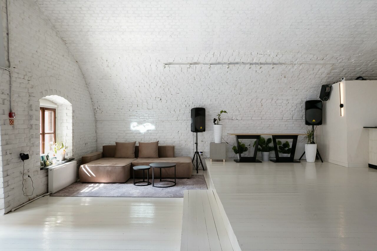 Арендовать Brodniki Loft (Бродники Лофт). Лофт из белого кирпича. Лофт-пространство с белыми кирпичными стенами и арочными потолками. Есть диван с подушками на ковре, два маленьких столика, растительность в белых вазах, акустические колонки, белые полы. Естественное освещение через окно.