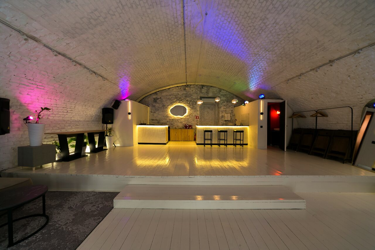 Арендовать Brodniki Loft (Бродники Лофт). Лофт из белого кирпича. Просторный подвал с арочным потолком из кирпича, пол из светлого дерева. Центральный бар с подсветкой и тремя стульями. На стенах декоративные светильники, разноцветная неоновая подсветка. Справа вешалка, слева кашпо с растениями.