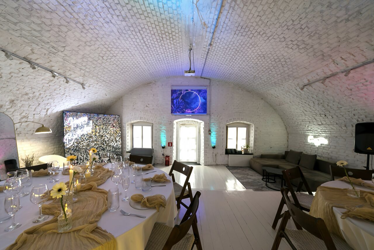 Арендовать Brodniki Loft (Бродники Лофт). Лофт из белого кирпича. Арочное помещение с белыми кирпичными стенами и потолком. В центре длинный стол с белой скатертью и посудой, украшенный цветами. Слева картина на мольберте, справа серый диван. Пол светлый. Освещение точечное и подвесное.