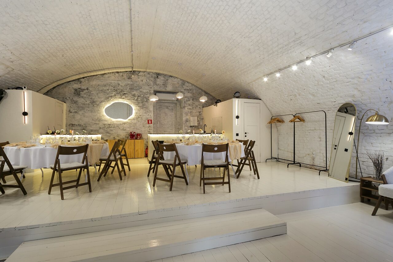 Арендовать Brodniki Loft (Бродники Лофт). Лофт из белого кирпича. Арочный зал с белыми кирпичными стенами. В центре установлены круглые столы с белыми скатертями и деревянными стульями. Небольшая сцена, деревянные полы, зеркало овальной формы. Освещение с помощью точечных светильников. Слева видны двери.