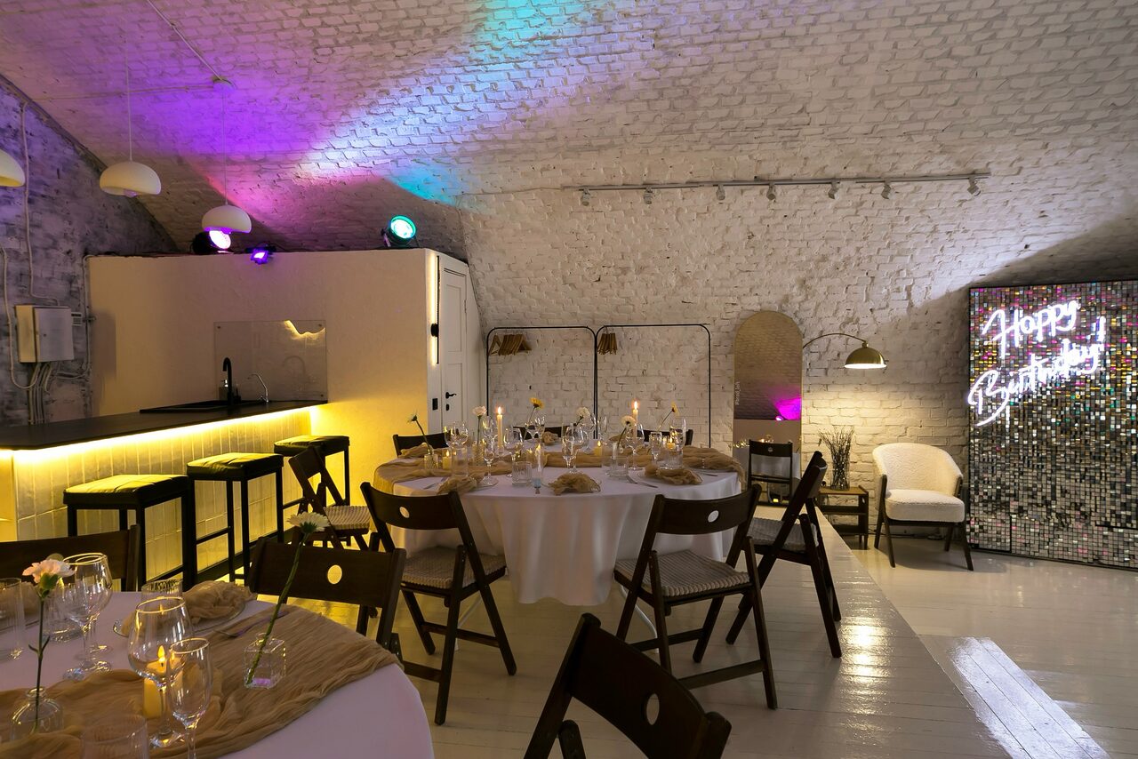 Арендовать Brodniki Loft (Бродники Лофт). Лофт из белого кирпича. Арендуемое пространство с кирпичными сводчатыми потолками, барной зоной с высокими стульями, круглыми столами с белыми скатертями и деревянными стульями. Декоративное освещение разноцветное, неоновая надпись "Happy Birthday", белые стены и пол.