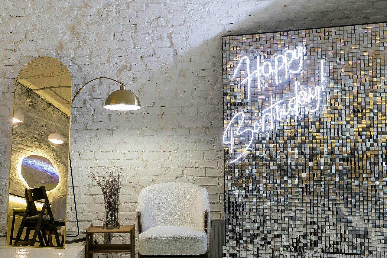 Арендовать Brodniki Loft (Бродники Лофт). Лофт из белого кирпича. Просторное помещение с кирпичными стенами, белое кресло, деревянный столик с вазой, настенное зеркало, стул. На стене неоновая надпись "Happy Birthday", плитка с зеркальной поверхностью. Арочный потолок с освещением в виде ламп.
