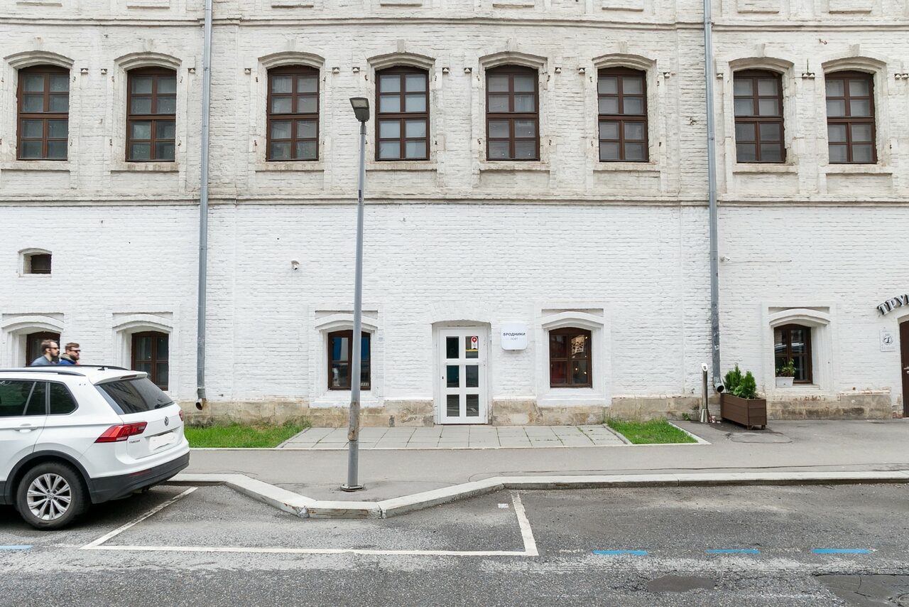 Арендовать Brodniki Loft (Бродники Лофт). Лофт из белого кирпича. Фасад кирпичного здания с большими окнами и центральной белой дверью. Перед зданием тротуар, фонарь и припаркованный белый автомобиль. Стены светлого цвета, на улице асфальтированная дорога и парковочные линии.