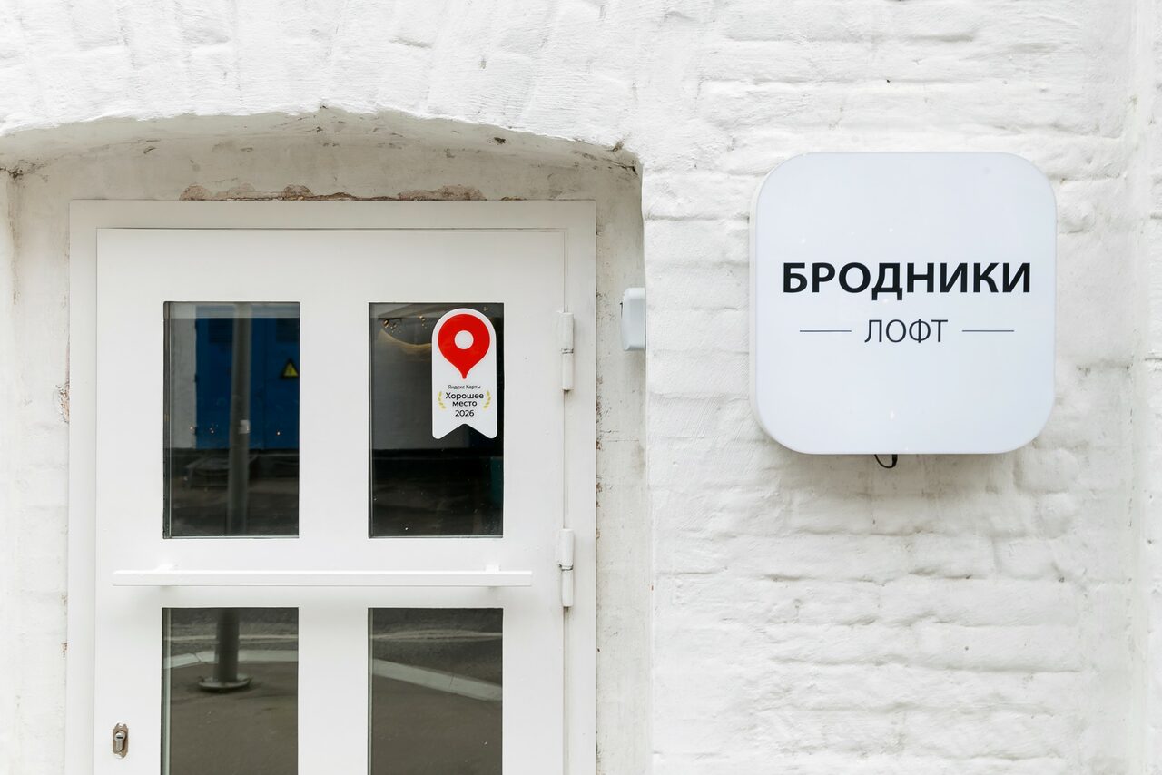Арендовать Brodniki Loft (Бродники Лофт). Лофт из белого кирпича. Вход с белой деревянной дверью с двумя стеклянными вставками и наклейкой с красным маркером. На белой кирпичной стене справа вывеска с текстом «Бродники Лофт».