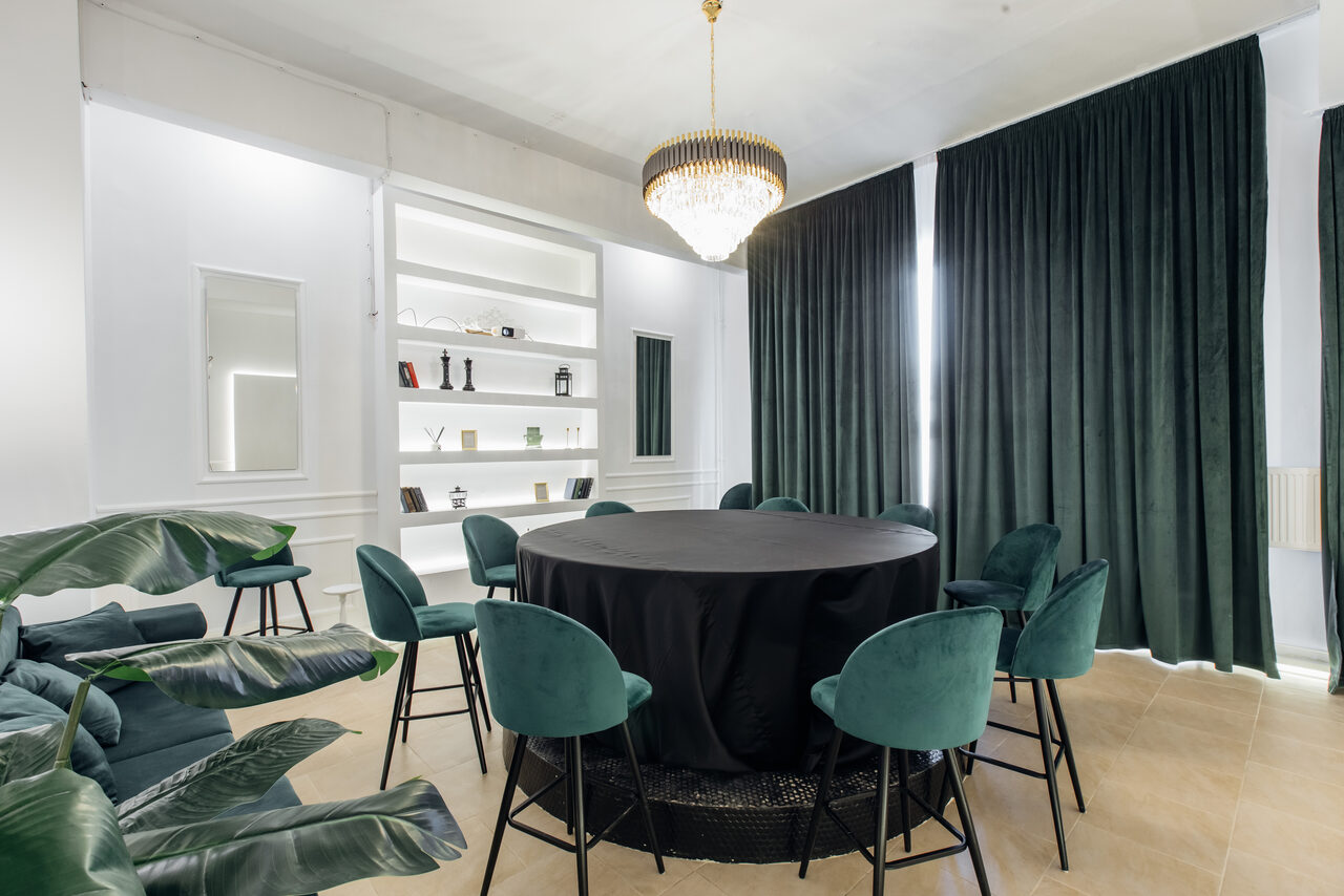 Арендовать Rubin Loft (Рубин Лофт). Зал Империал. Комната с круглым столом, покрытым черной скатертью, окруженным восемью зелеными стульями. На стене стеллаж с книгами и декором. Большие темные шторы, светлый пол. Центральная люстра. Зеленые растения в углу.