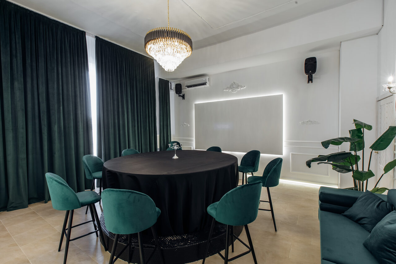 Арендовать Rubin Loft (Рубин Лофт). Зал Империал. Современное помещение с круглым столом, покрытым черной скатертью, девятью стульями с зелеными сиденьями, люстрой, зелеными шторами, кондиционером, светлым деревянным полом, декоративными элементами на стене, растением в горшке и нейтральным освещением.