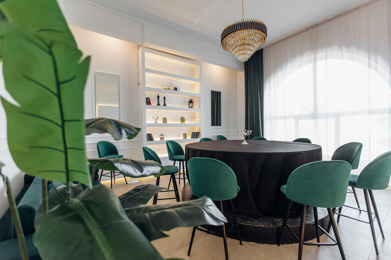 Арендовать Rubin Loft (Рубин Лофт). Зал Империал. Современное помещение с круглым столом, окружённым зелёными стульями. Наземные растения на переднем плане. На заднем плане встроенные полки с декором. Большое окно с прозрачными шторами. Подвесная люстра. Светлые стены и пол.