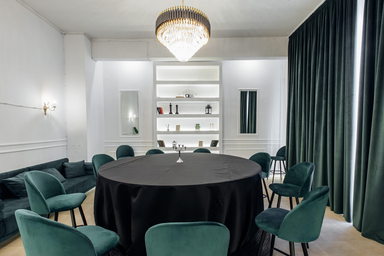 Арендовать Rubin Loft (Рубин Лофт). Зал Империал. Круглый стол с черной скатертью окружен бирюзовыми стульями. Справа зеленые шторы, слева диван. На стене встроенные полки с предметами, освещенные сверху. Центральное освещение — люстра. Цвета: белый, бирюзовый, зеленый, черный.