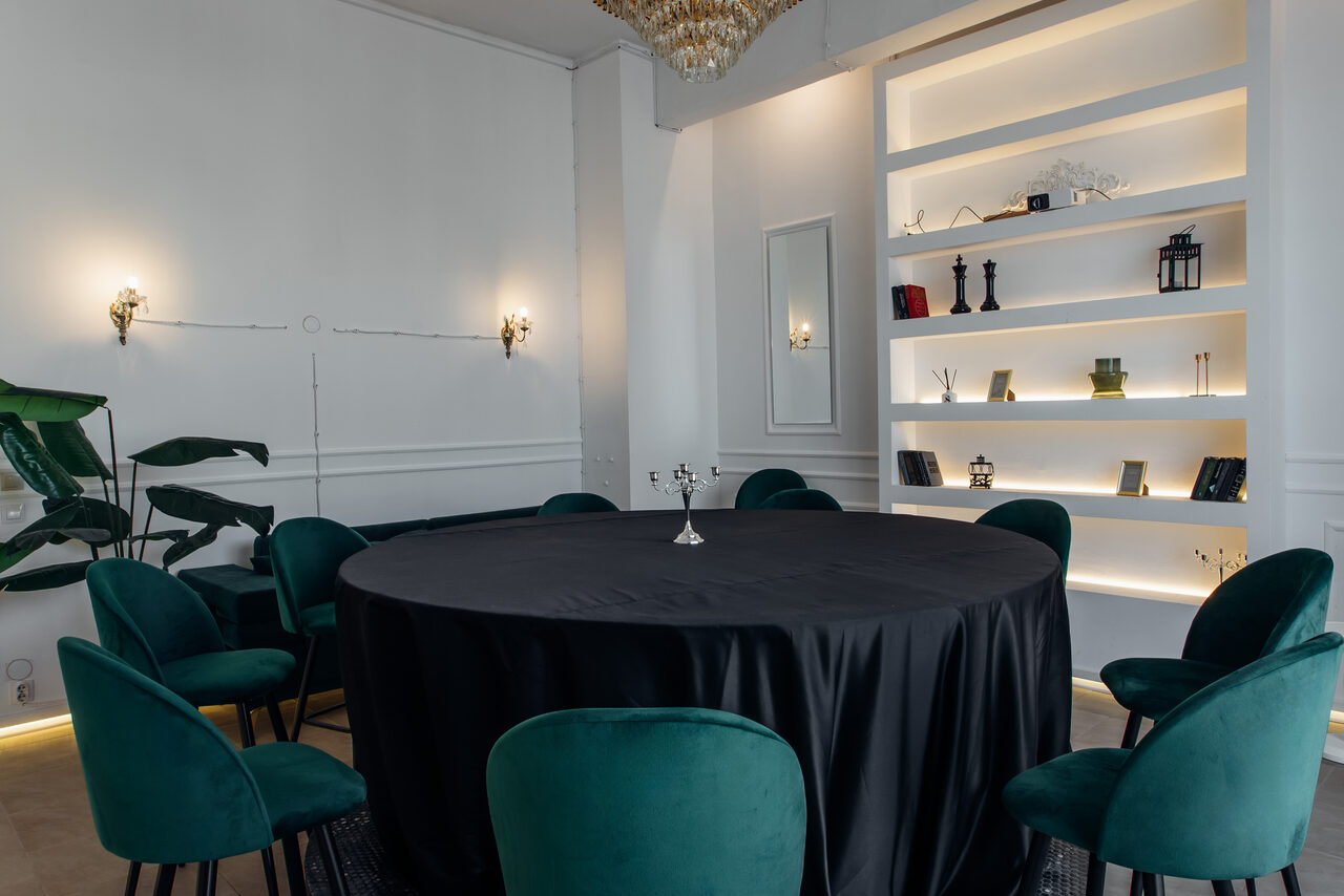 Арендовать Rubin Loft (Рубин Лофт). Зал Империал. Пространство с круглым столом, покрытым черной скатертью, окруженным зелеными стульями. На стене подсвеченные полки с декором, два бра и зеркало. Потолок с люстрой, белые стены, паркет. Зеленое растение сбоку.