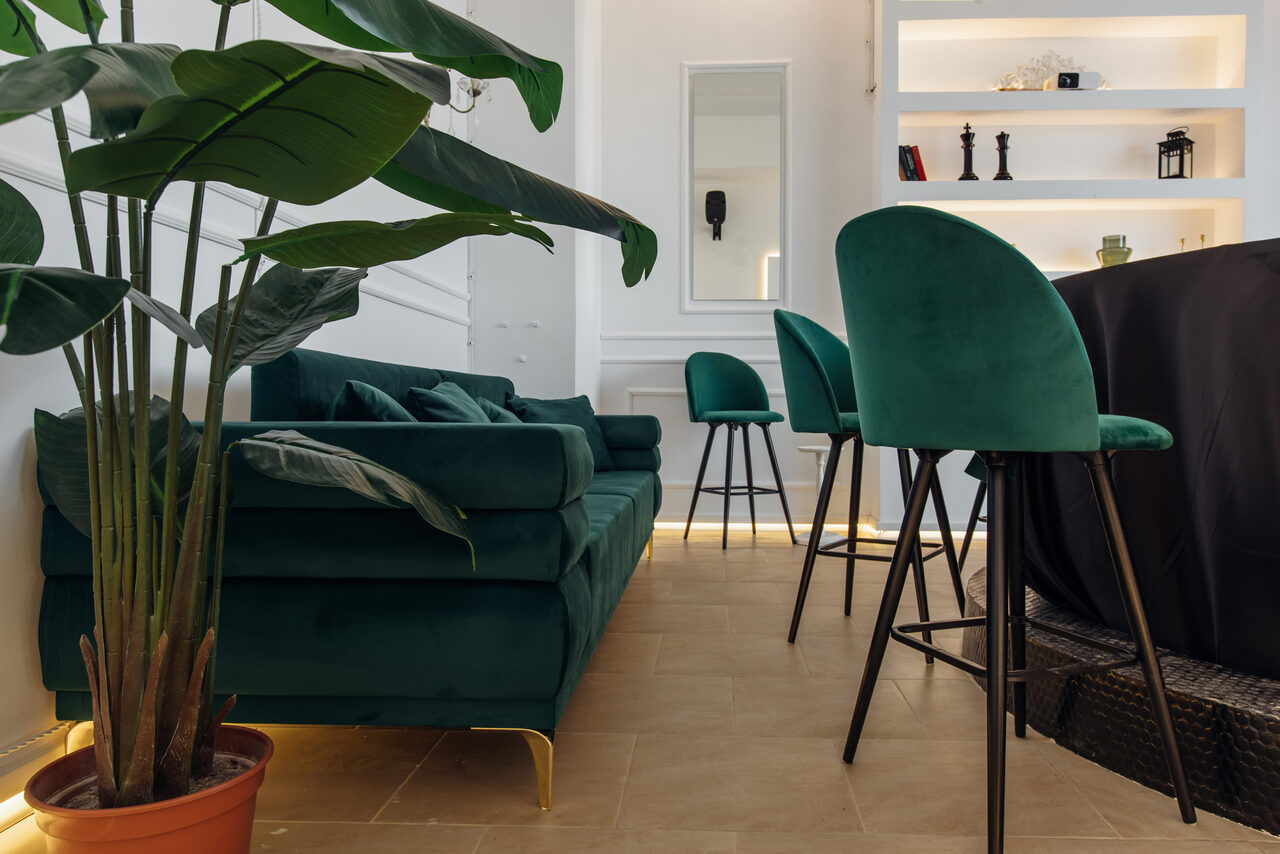 Арендовать Rubin Loft (Рубин Лофт). Зал Империал. Современное пространство с зелёным диваном и барными стульями, расположенными у стойки. Комната с деревянным полом, белыми стенами и встроенными полками с декором. Растение в горшке, мягкое освещение, зеркало на стене.